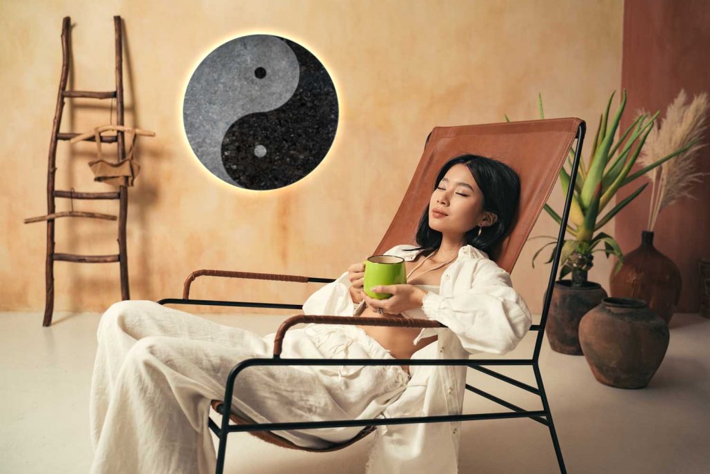 Asiatische Frau sitzt mit einer grünen Tasse auf einem Liegestuhl vor runder Infrarotheizung Natursteinheizung Yin und Yang mit Hintergrundbeleuchtung.